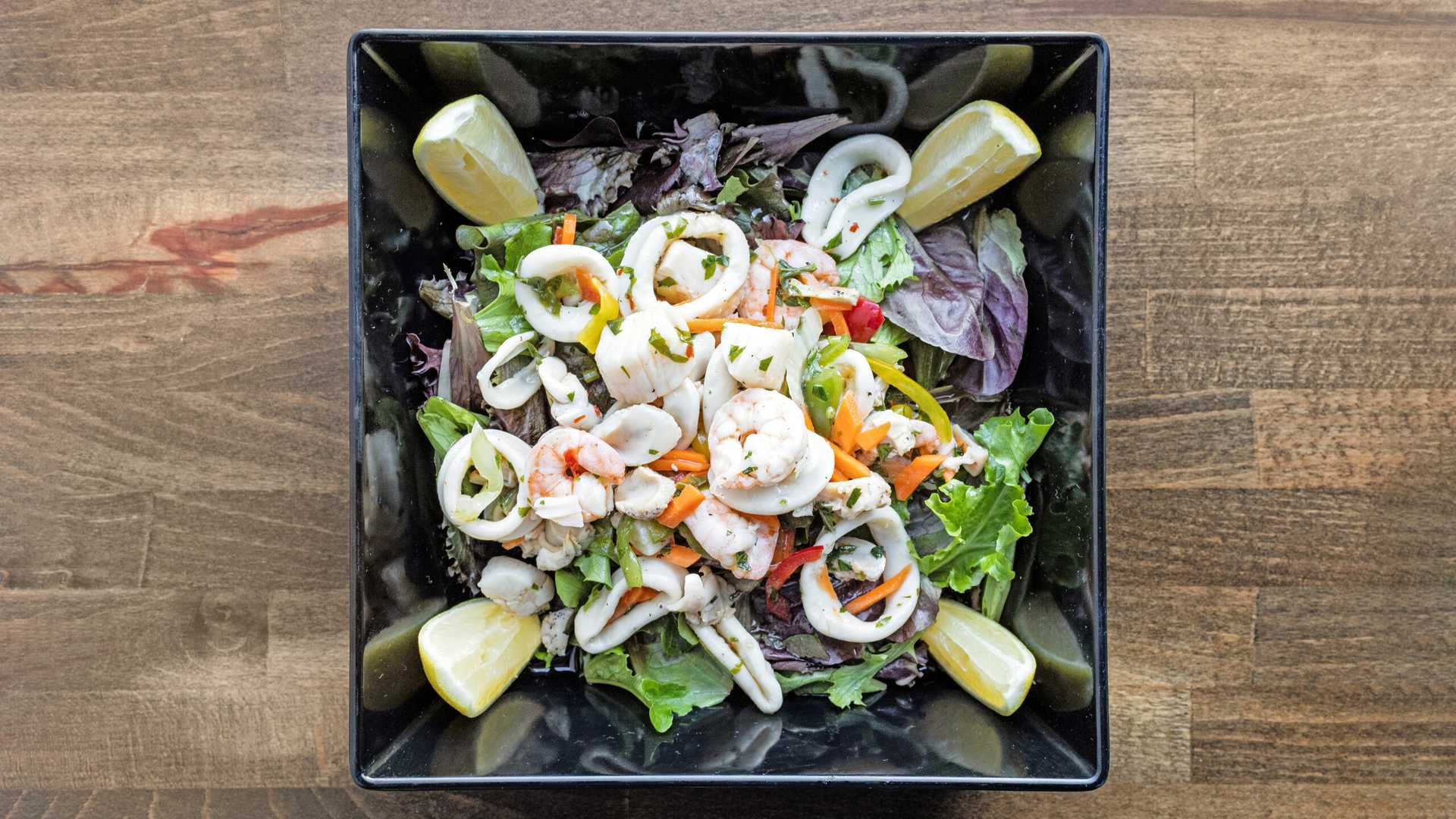 Seafood salad with shrimp, calamari rings, mixed greens, sliced vegetables, and lemon wedges in a black square bowl on a wooden surface.