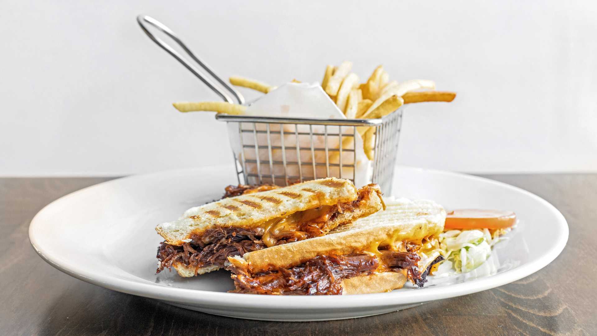 Short rib grilled cheese panini sandwich with melted cheese served on a white plate accompanied by French fries in a small metal basket and a side of shredded lettuce and tomato slice.
