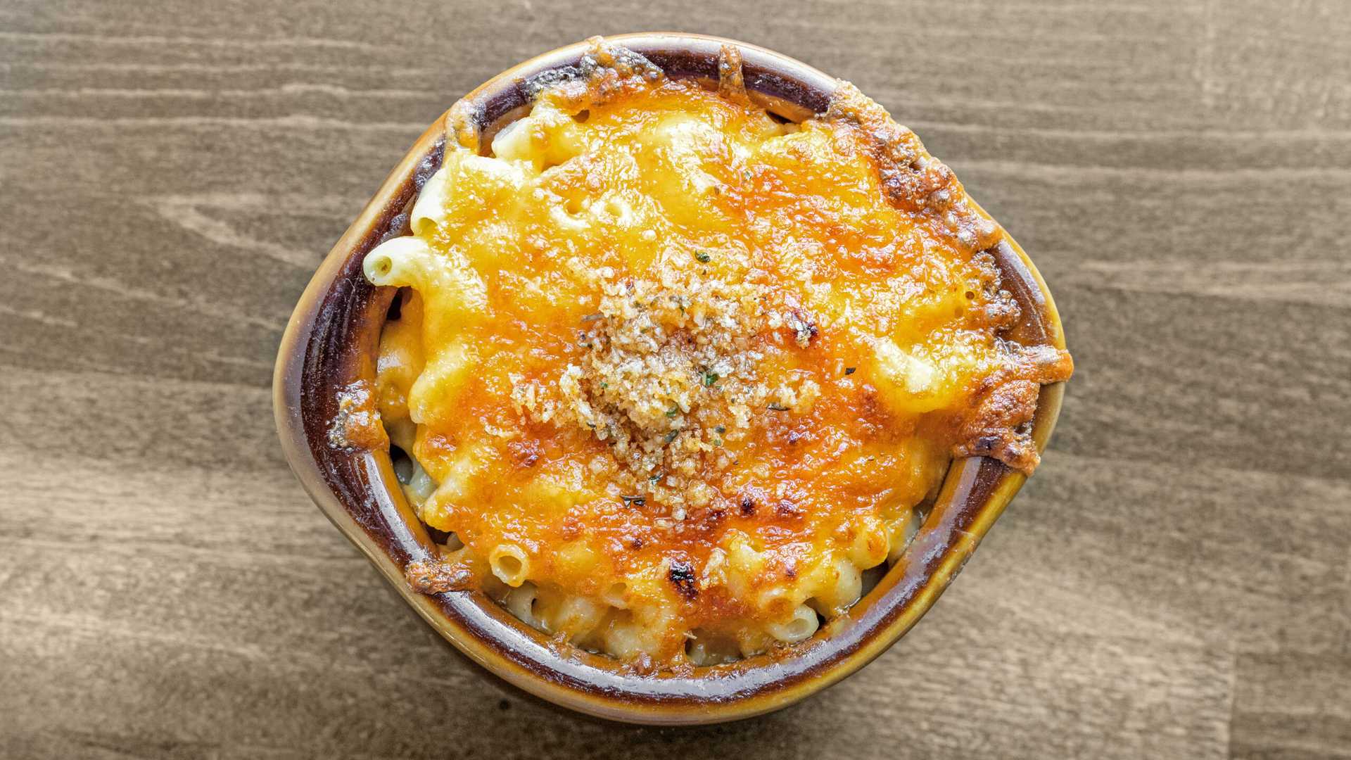 Baked truffle mac and cheese topped with melted golden cheese and breadcrumbs in a brown ceramic bowl on a wooden surface.