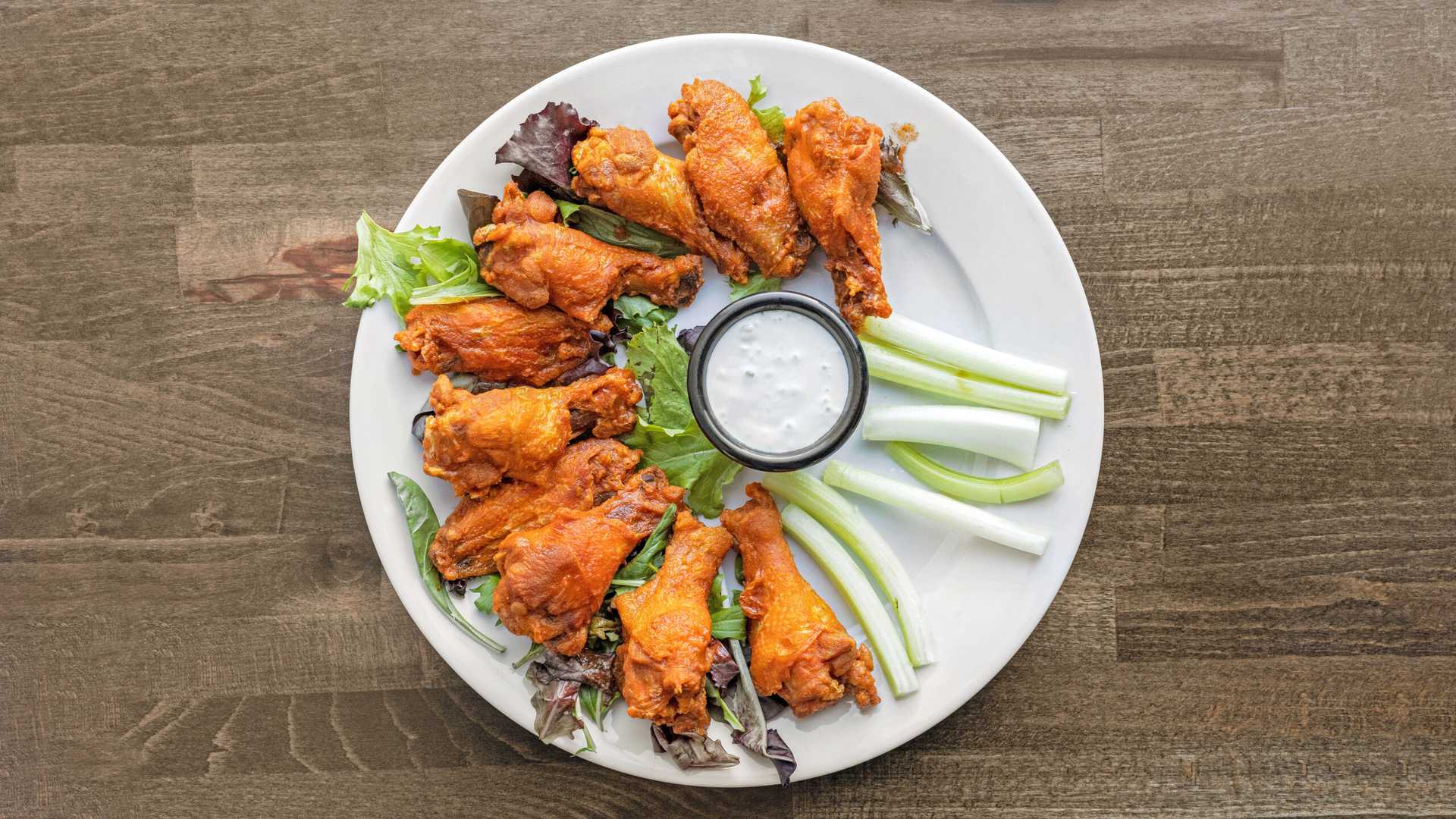 Plate of buffalo chicken wings arranged around a cup of ranch dressing, garnished with lettuce and celery sticks on a wooden table.