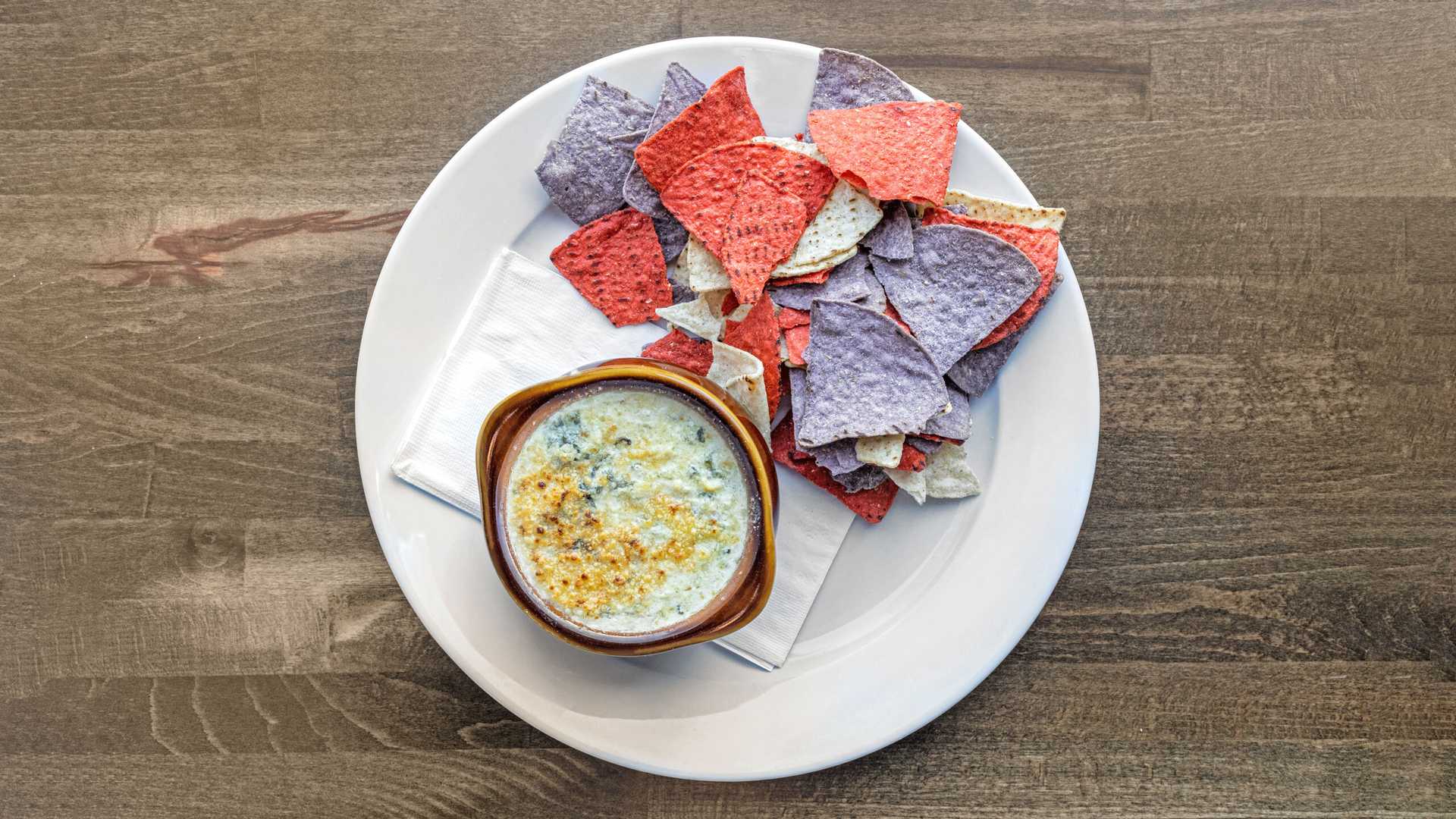 White plate with a bowl of creamy spinach and artichoke dip next to multicolored red, white, and blue tortilla chips on a wooden table.