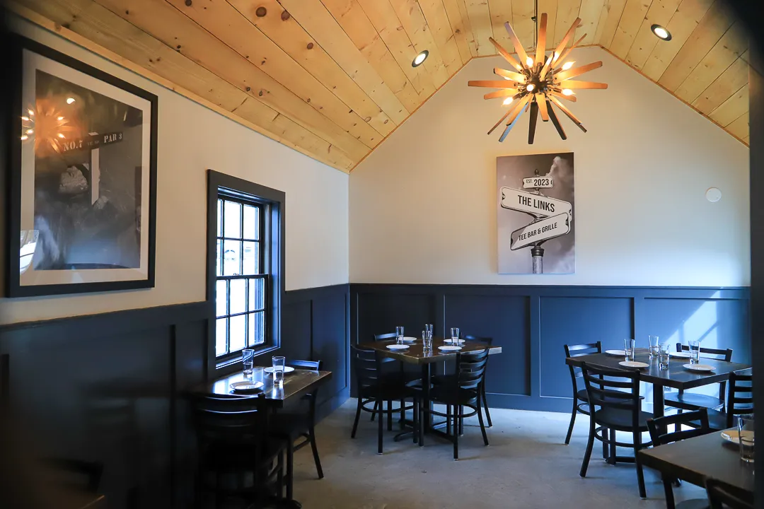 Cozy dining area with dark wood tables and chairs, wooden ceiling, and wall art featuring a sign for 'The Links Tee Bar & Grille'.