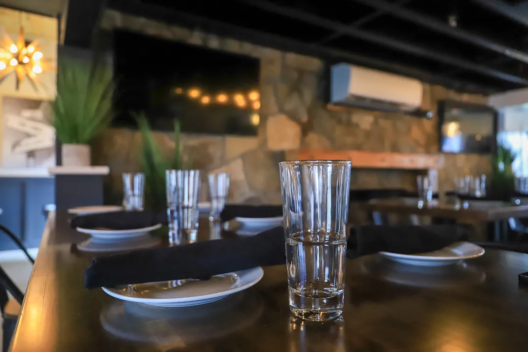 Close-up of a glass of water on a dark wooden table set with plates and black napkins in a cozy restaurant interior with stone walls and greenery.