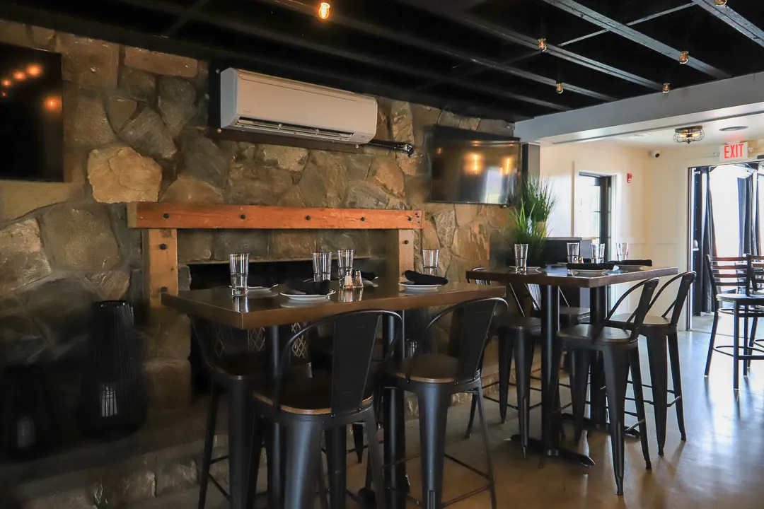 Dining area with two tall wooden tables, black metal chairs, stone wall, mounted air conditioner, and two wall-mounted TVs.