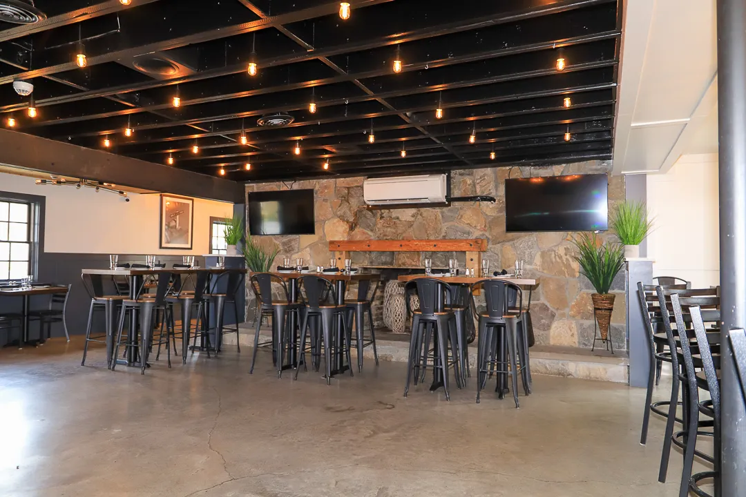 Modern restaurant interior with high black metal chairs, wooden tables, stone wall with mounted TVs, and hanging string lights on a black ceiling.