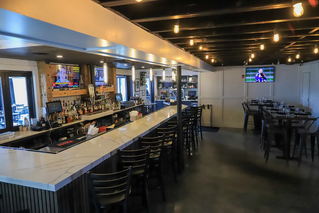 Interior of a modern bar with a long white marble counter, multiple chairs, bottles behind the bar, and three TVs on the walls.