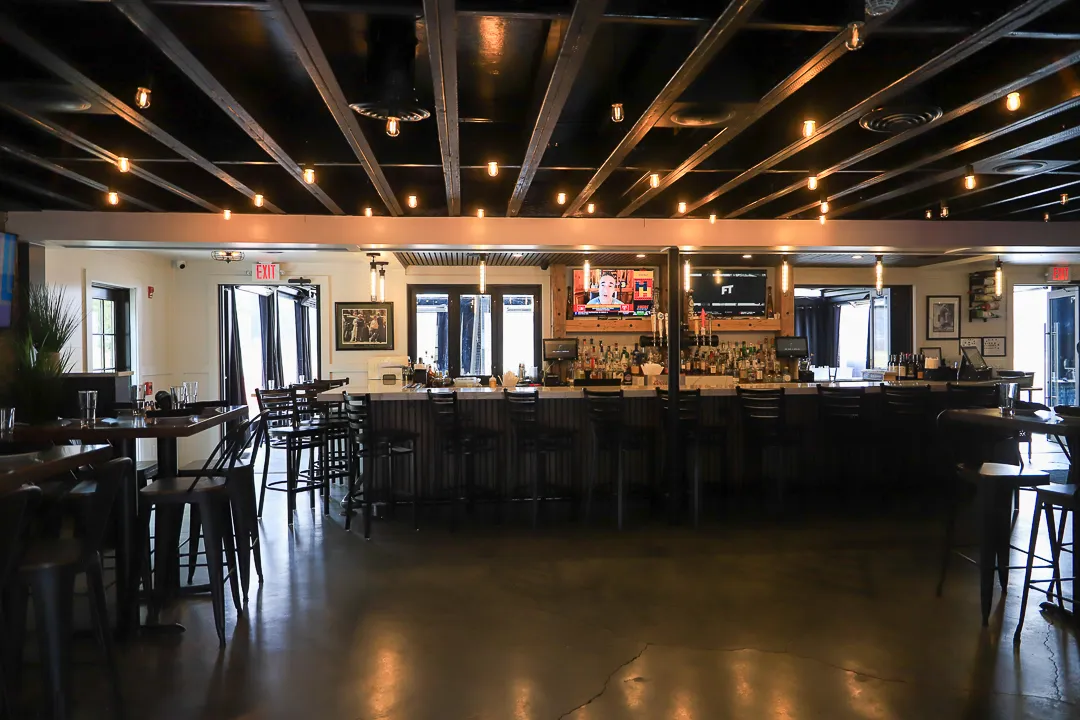 Interior of a stylish bar with a long counter, bar stools, hanging lights on a black ceiling, and two TVs mounted behind the bar.