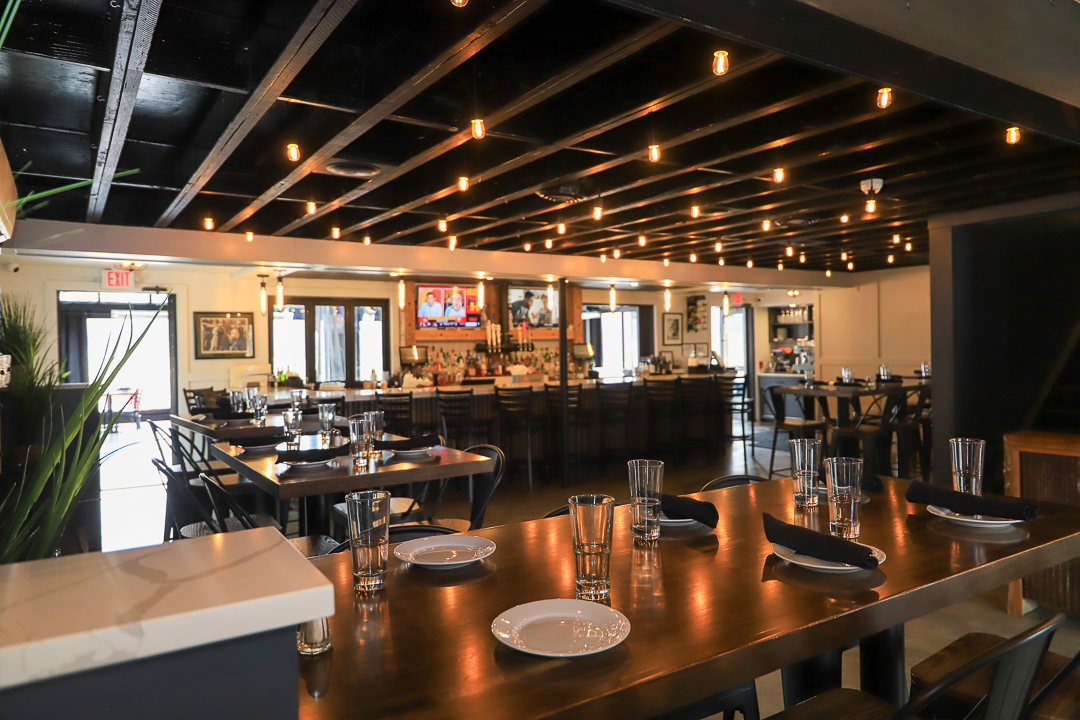 Cozy restaurant interior with wooden tables set with plates, glasses, and black napkins under warm hanging lights and a bar with stools in the background.