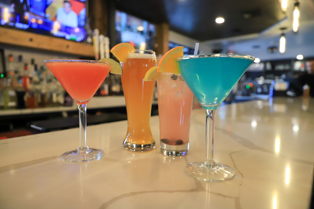 Four colorful cocktails with fruit garnishes on a bar counter in a dimly lit bar.