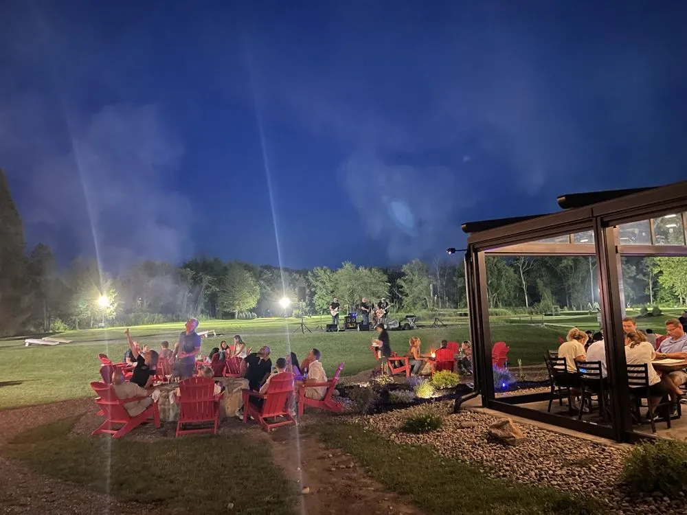 People gathered outdoors at night sitting around a fire pit in red chairs with a band playing in the background on a grassy field.
