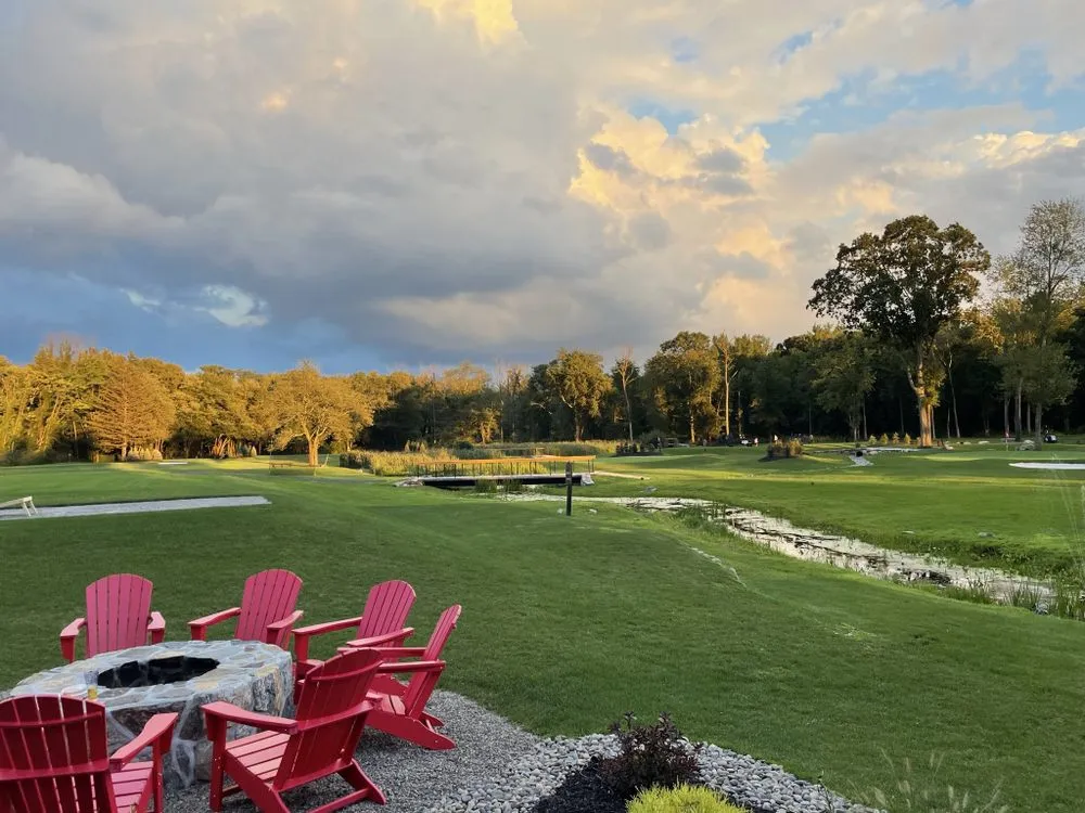 Red Adirondack chairs arranged around a stone fire pit on a grassy golf course under a partly cloudy sky at sunset.