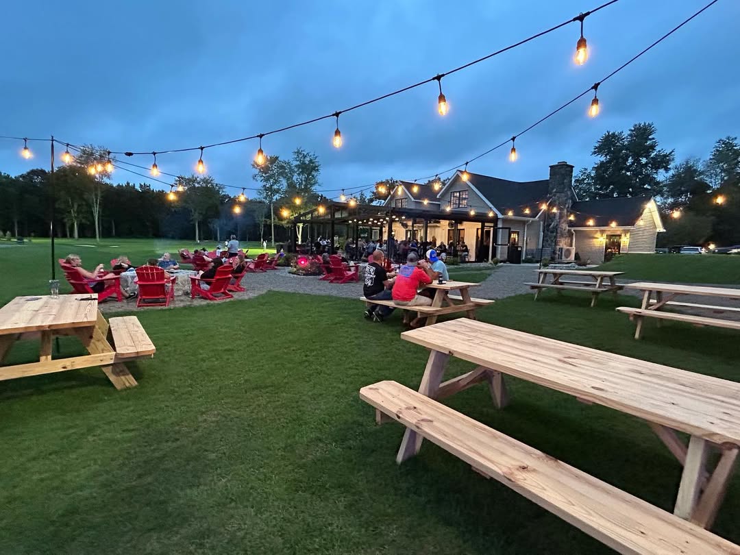 Evening outdoor seating area with string lights, red Adirondack chairs around fire pits, picnic tables, and a building with people inside and outside.