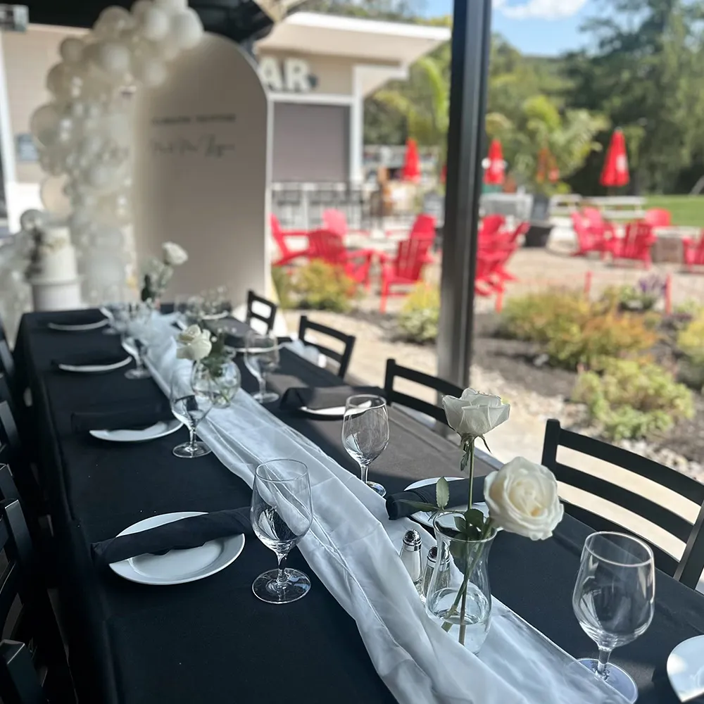 Elegant dining table with black tablecloth and white runner, set with white roses, glassware, and black napkins in a bright room overlooking a garden patio with red chairs.