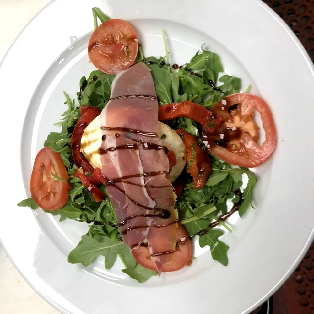 Plate with arugula salad topped with sliced tomatoes, roasted red peppers, burrata cheese, prosciutto, and balsamic glaze drizzle.