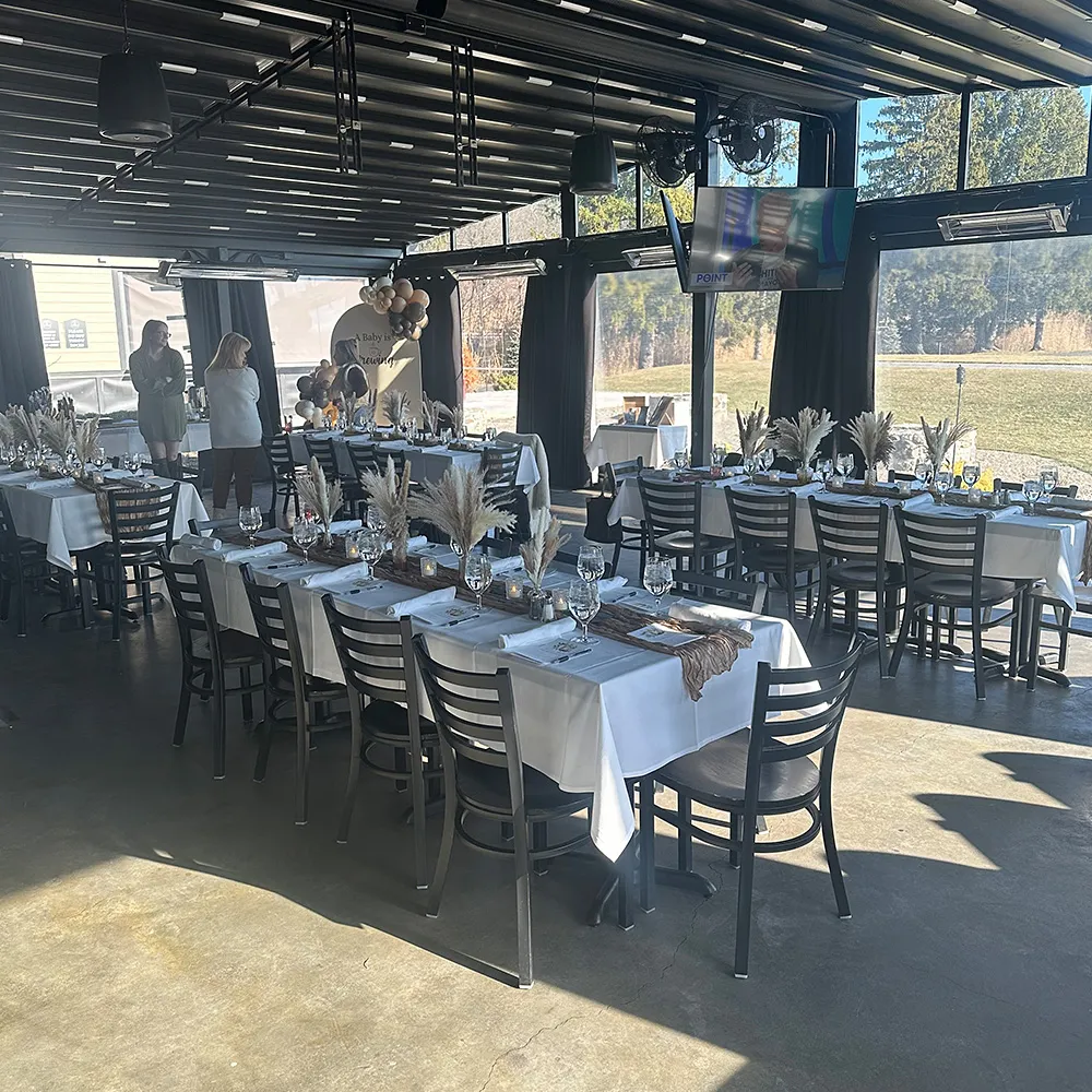 Indoor event space set up with long tables covered in white tablecloths and decorated with pampas grass centerpieces, with three people standing near a balloon arch in the background.