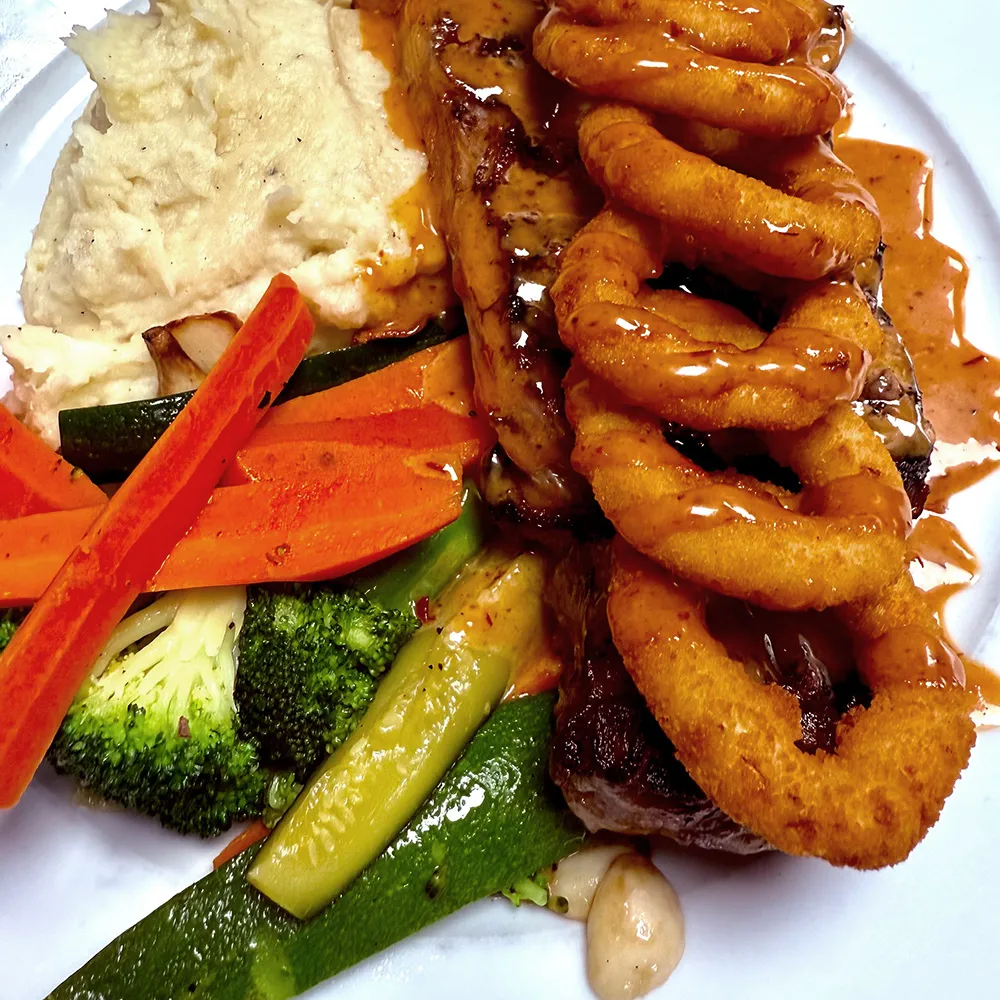 Plate with mashed potatoes, grilled vegetables, steak topped with fried onion rings and brown gravy.