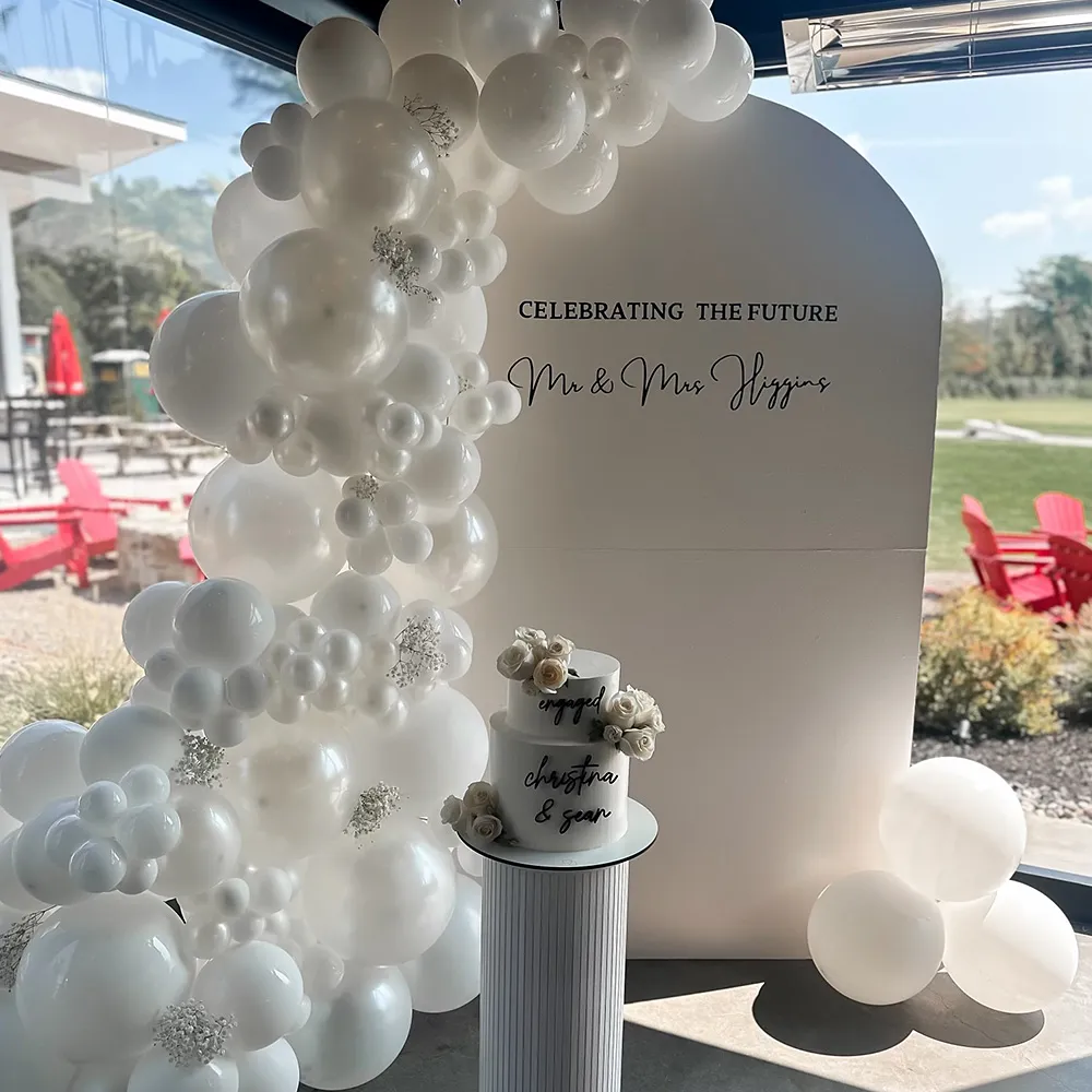 White balloon arch and two-tier engagement cake with flowers next to a backdrop reading 'Celebrating the Future Mr & Mrs Higgins.'