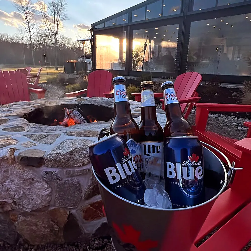 Bucket of beers on a stone table near a lit outdoor fire pit with red chairs and a building at sunset.
