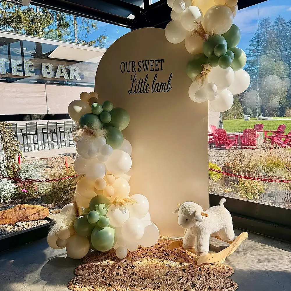 Decorative setup with green and white balloons around a beige backdrop reading 'Our Sweet Little Lamb' and a plush rocking lamb on a woven rug.
