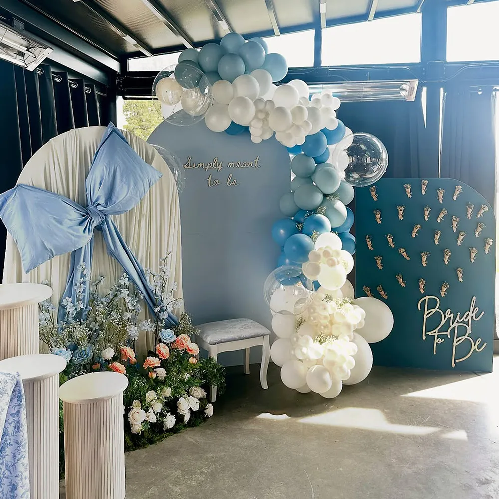Decorative bridal shower setup with white and blue balloon arch, large blue bow, floral arrangements, and signs reading 'Simply meant to be' and 'Bride to Be'.