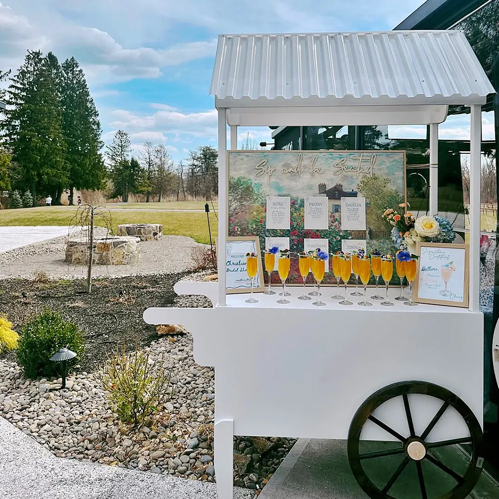 White drink cart with a roof, holding ten orange cocktails garnished with purple flowers, in a garden setting under a partly cloudy sky.