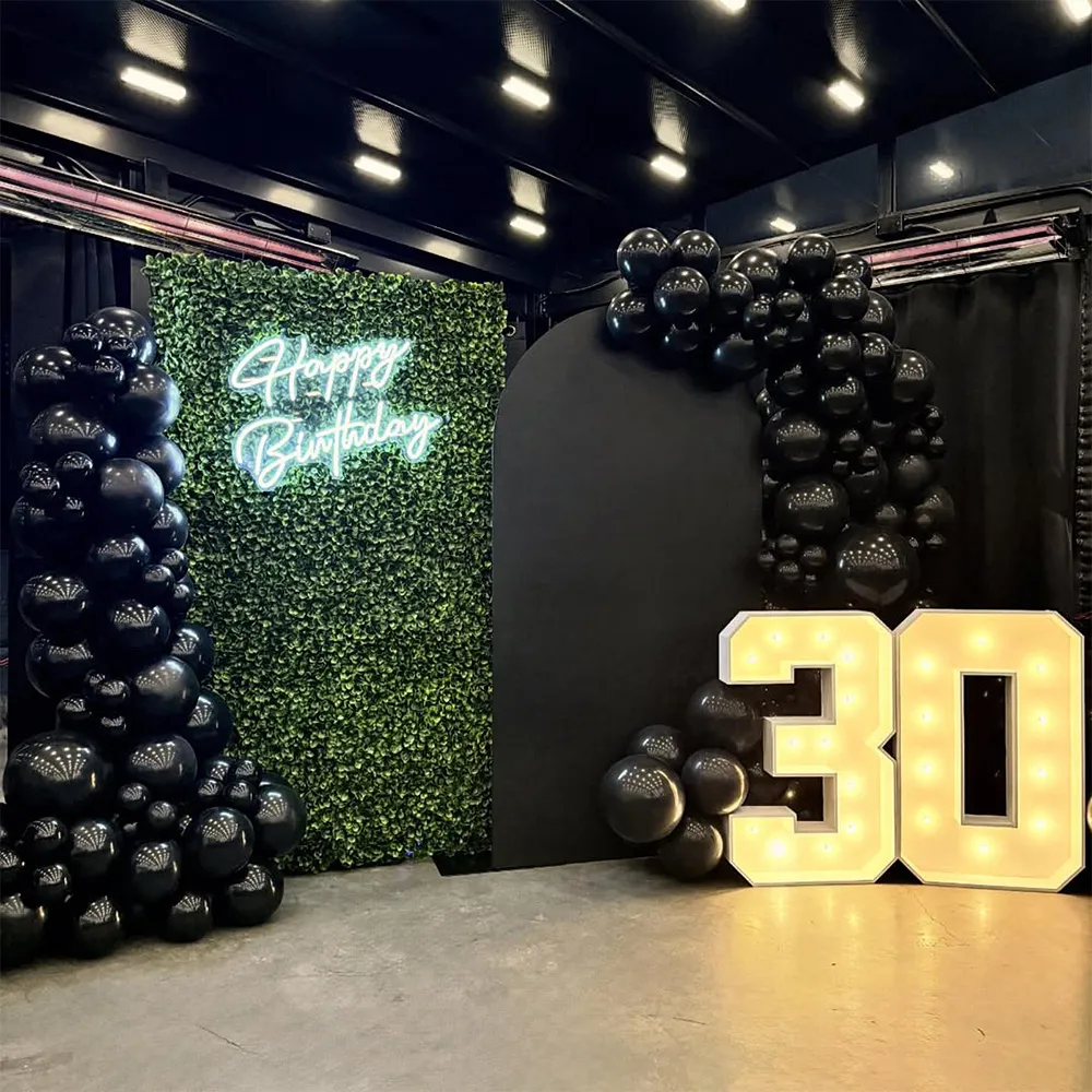 Birthday party setup with a green leafy backdrop featuring a neon 'Happy Birthday' sign, large illuminated '30' numbers, and black balloon decorations.