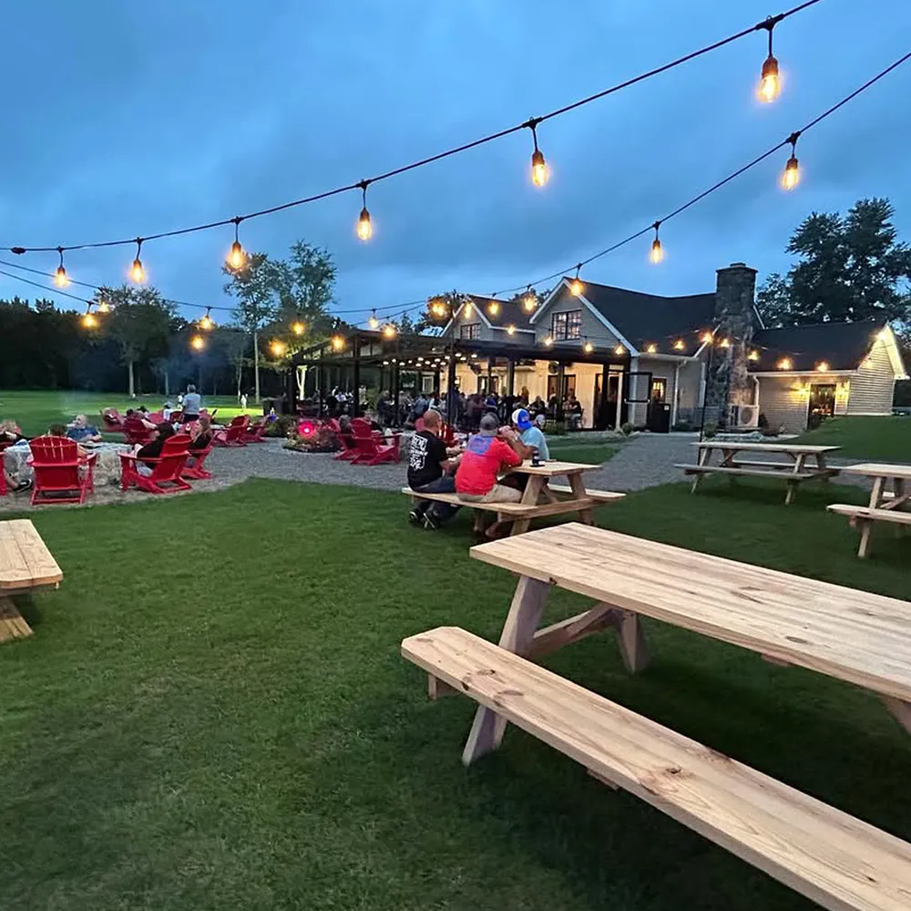 Outdoor social gathering at dusk with string lights over picnic tables and red chairs near a house with a lit porch.