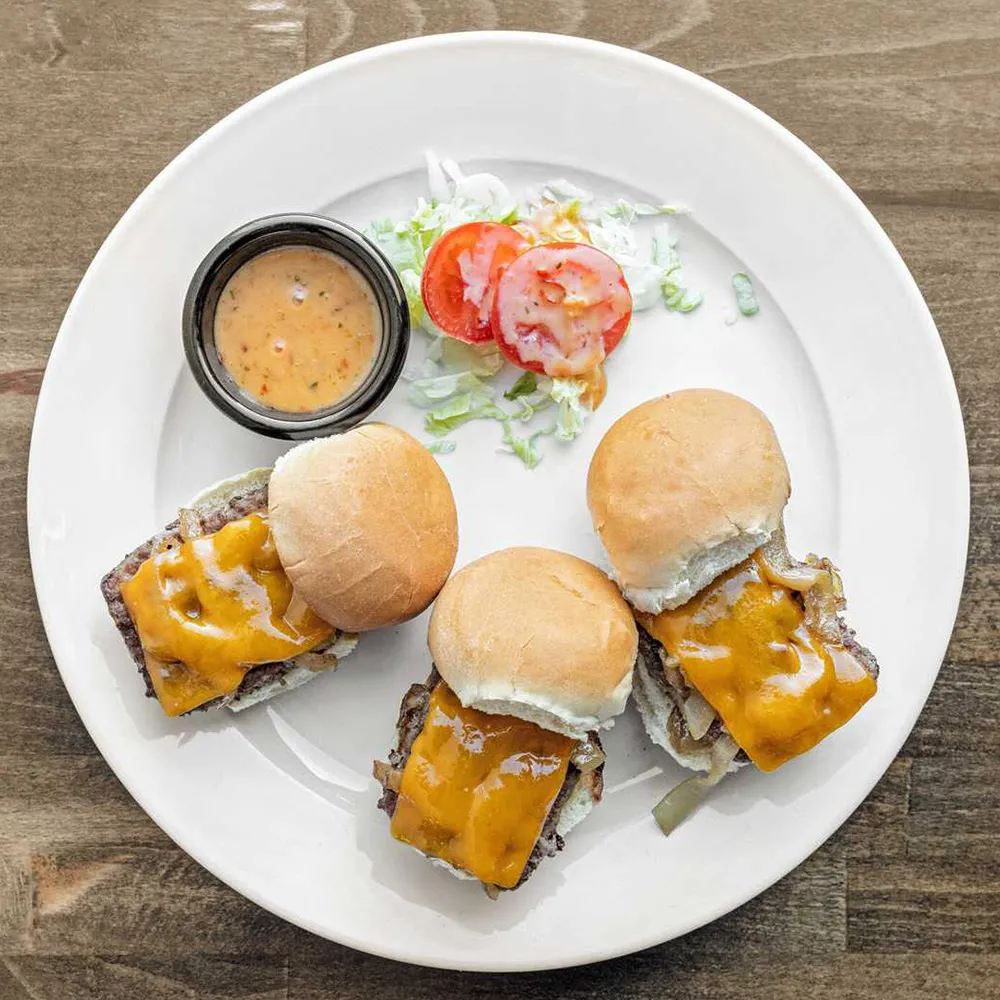 Plate with three cheeseburger sliders topped with melted cheddar, tomato slices, shredded lettuce, and a side of dipping sauce.