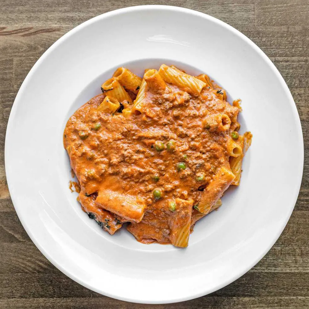 White plate with rigatoni pasta covered in a creamy meat and tomato sauce with peas, placed on a wooden surface.