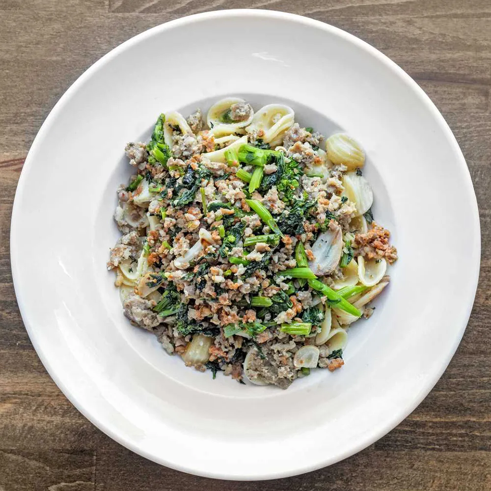 White plate with orecchiette pasta topped with ground meat and sautéed greens on a wooden surface.