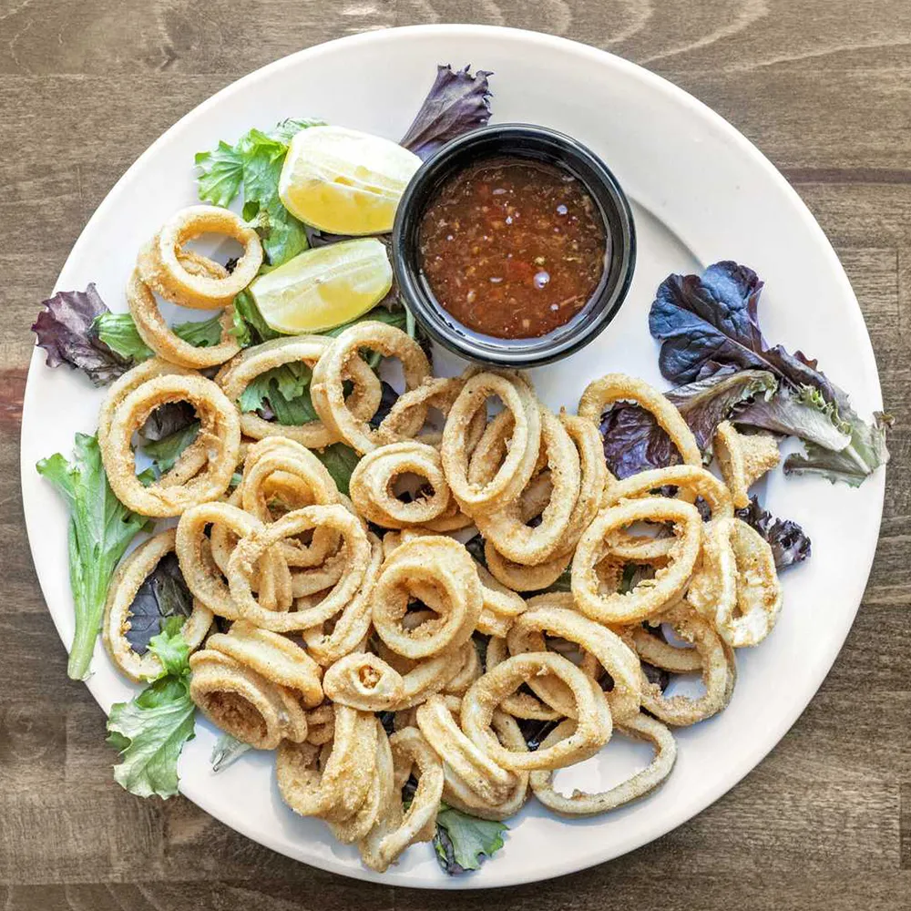 Plate of fried calamari rings served on mixed greens with lemon wedges and a container of dipping sauce.
