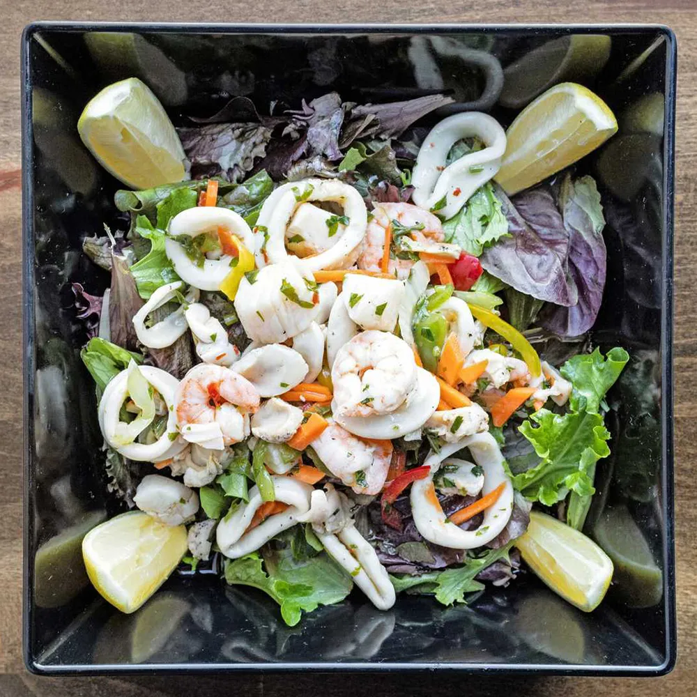 Seafood salad with shrimp, calamari rings, mixed greens, julienned vegetables, and lemon wedges in a black square bowl.