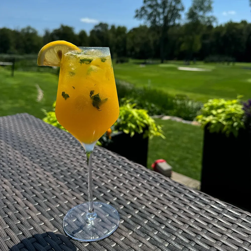 Yellow-orange cocktail with mint leaves and a lemon wedge garnish on a wicker table with a green outdoor landscape in the background.