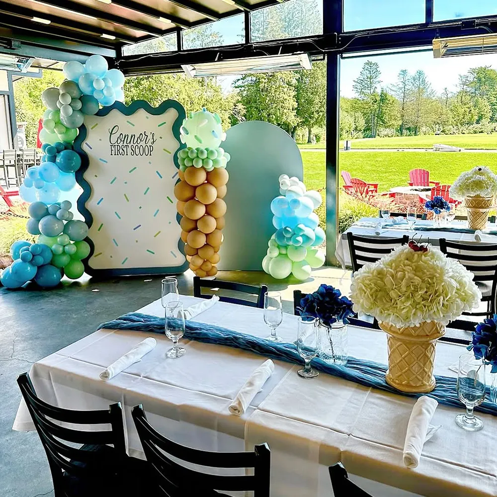Indoor party setup with tables covered in white cloth, decorated with blue and white ice cream cone flower centerpieces and balloon arrangements near a sign reading 'Connor's First Scoop'.