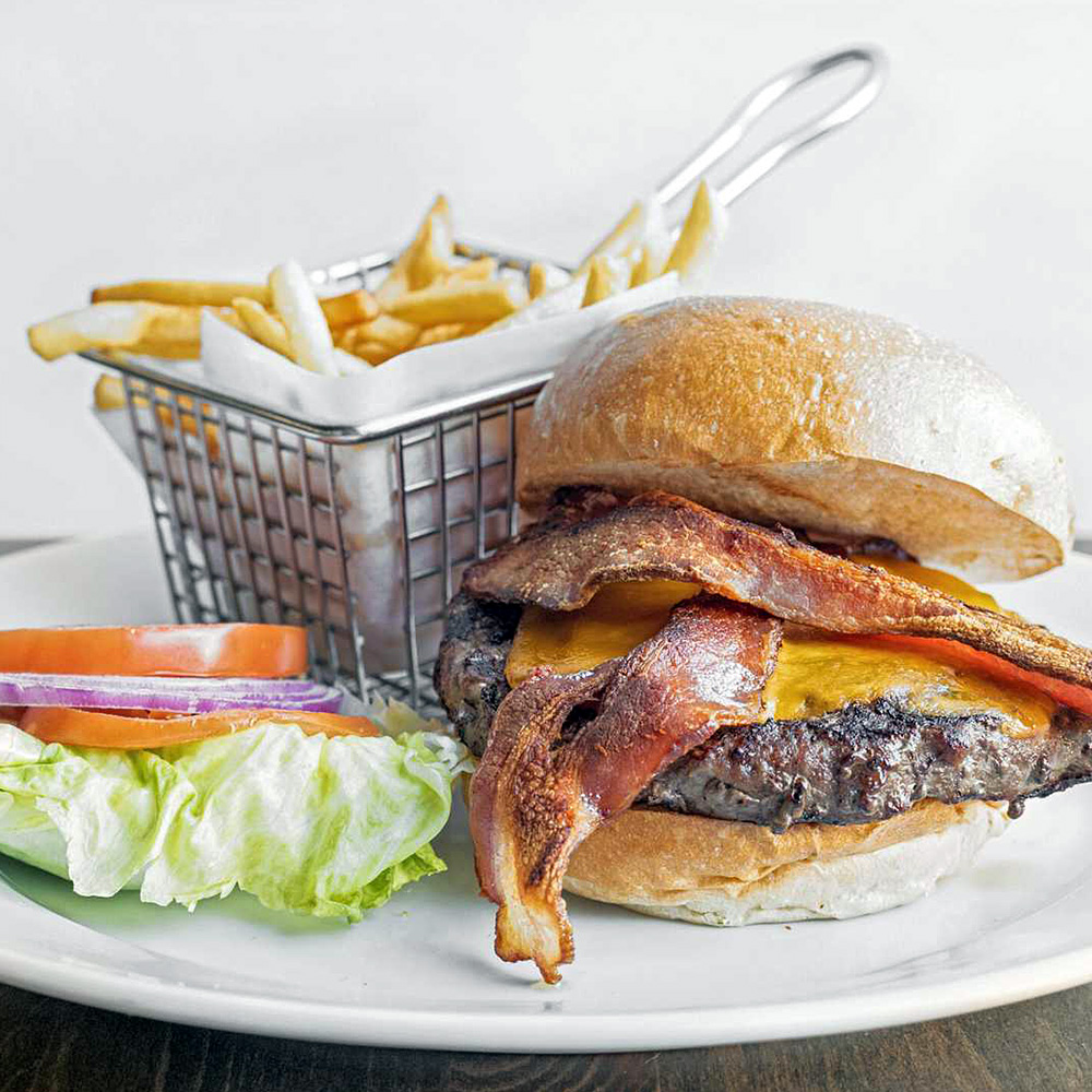 Bacon cheeseburger with lettuce, tomato, and onion on a plate next to a small basket of French fries.