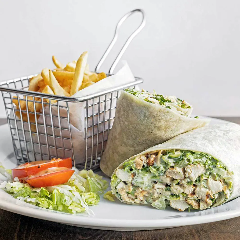Grilled chicken Caesar wrap cut in half served with French fries and a small side of lettuce and tomato on a white plate.