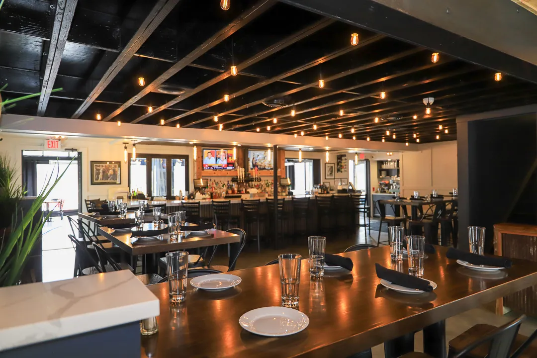 Modern restaurant interior with wooden tables set with plates, glasses, and black napkins under a ceiling with exposed beams and hanging lights.