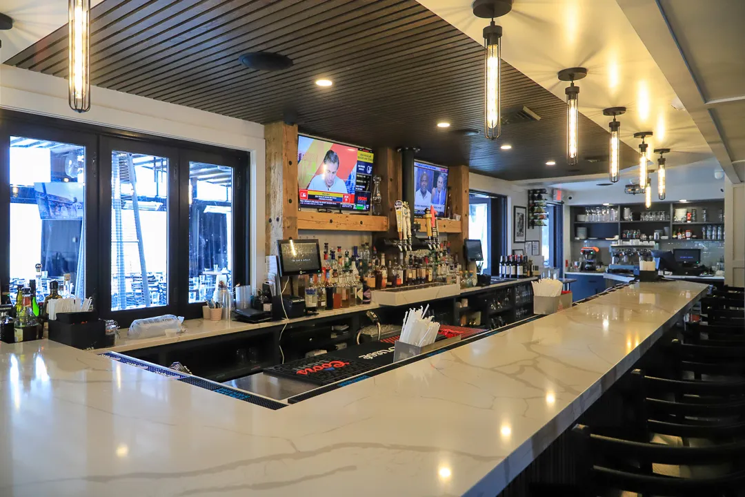 Interior view of a modern bar with marble countertops, multiple liquor bottles, mounted TVs showing news, and hanging pendant lights.