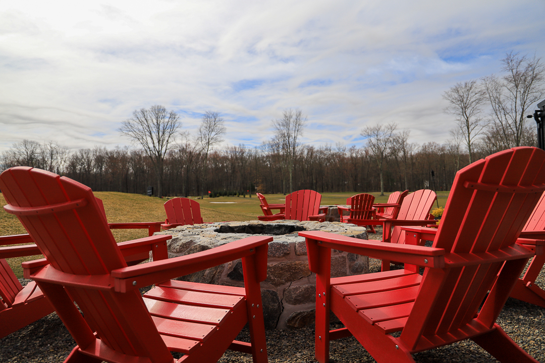 Circular outdoor stone fire pit surrounded by several red Adirondack chairs on a gravel surface with a grassy field and bare trees in the background under a partly cloudy sky.