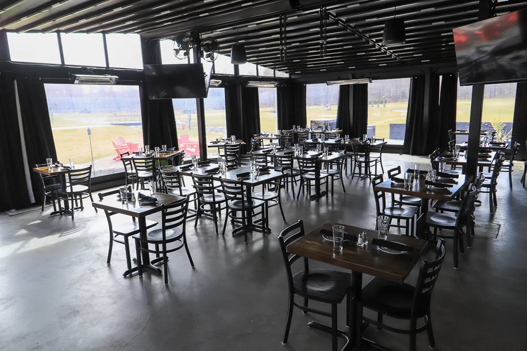 Empty modern restaurant interior with multiple tables and chairs set with glassware and napkins, large windows showing outdoor seating and grassy area.
