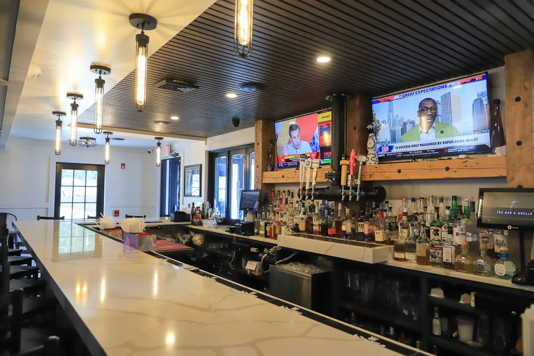Modern bar with white marble counter, hanging pendant lights, two TVs showing sports, and a variety of liquor bottles behind the bar.