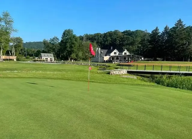 Golf green with a red flag, a wooden bridge, a clubhouse, and surrounding trees under a clear blue sky.