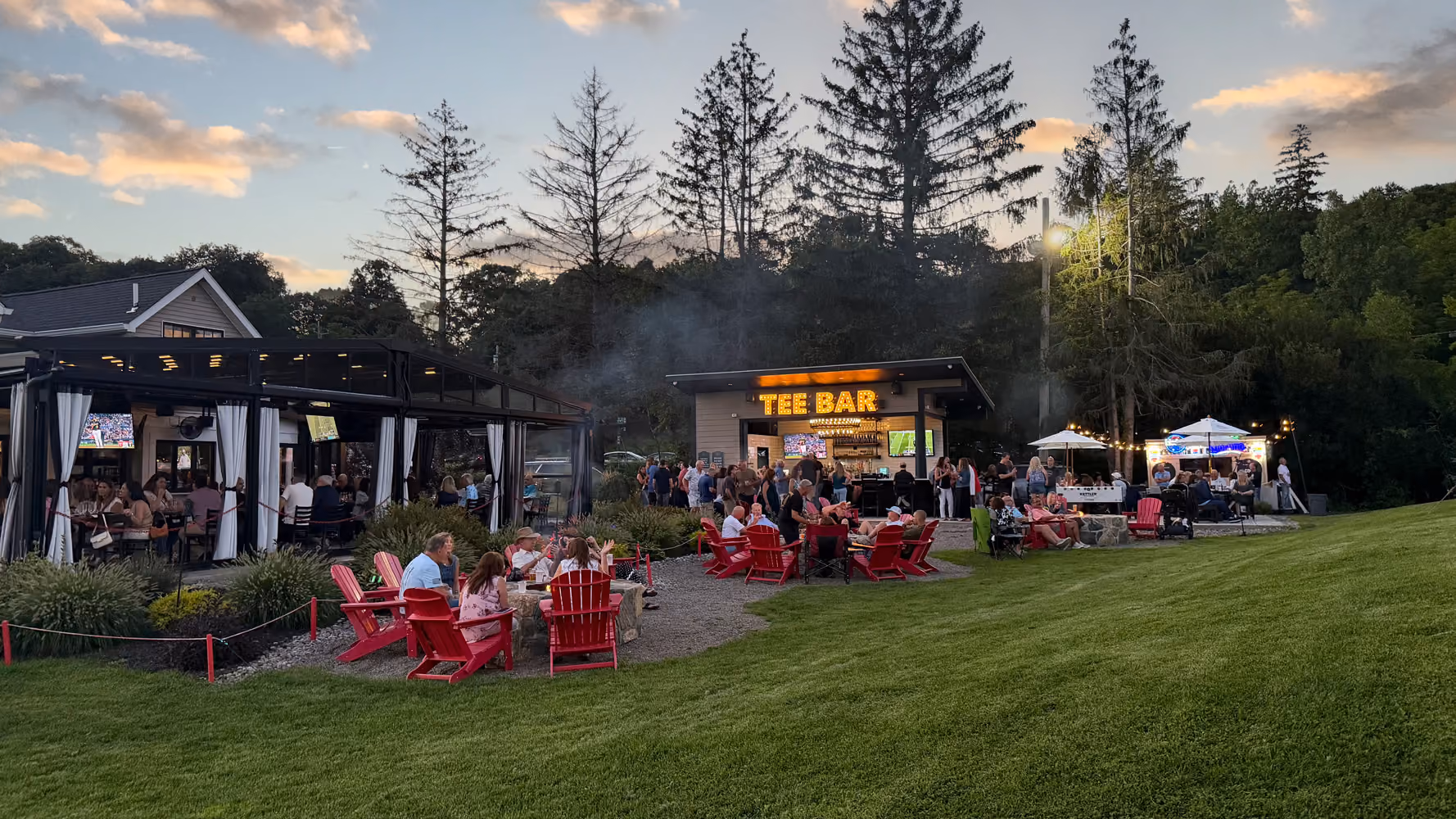 People sitting in red chairs around fire pits and gathering near a bar labeled Tee Bar at dusk outdoors.