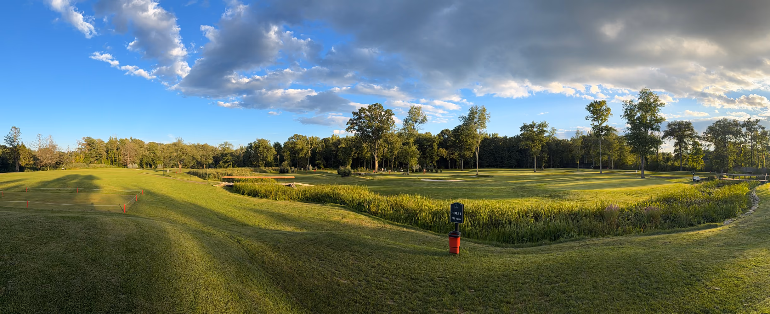 Wide view of a golf course with green fairways, trees, blue sky, and scattered clouds at sunset.