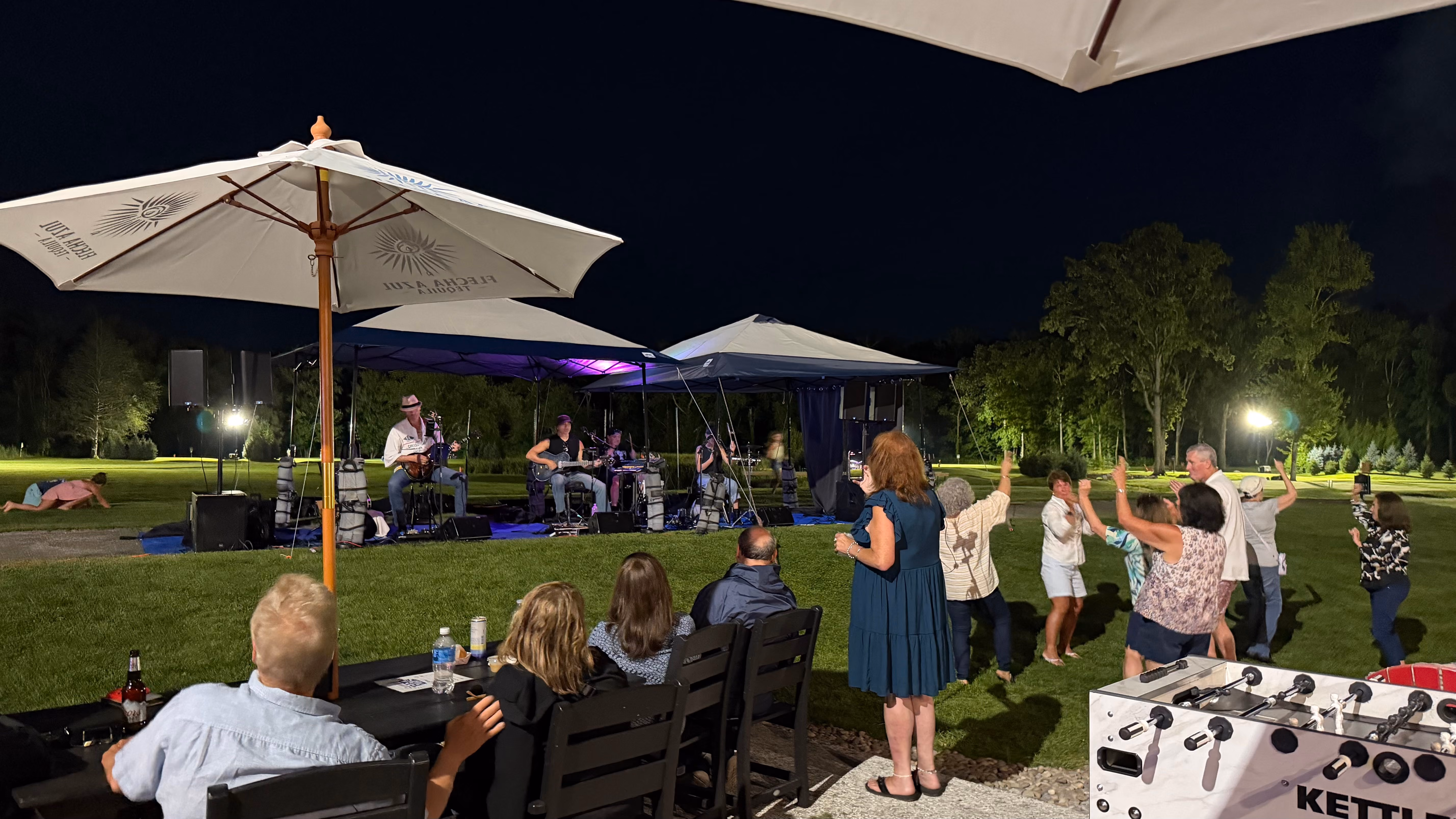 Outdoor night concert with a band on stage and people dancing and sitting on grass under large umbrellas.