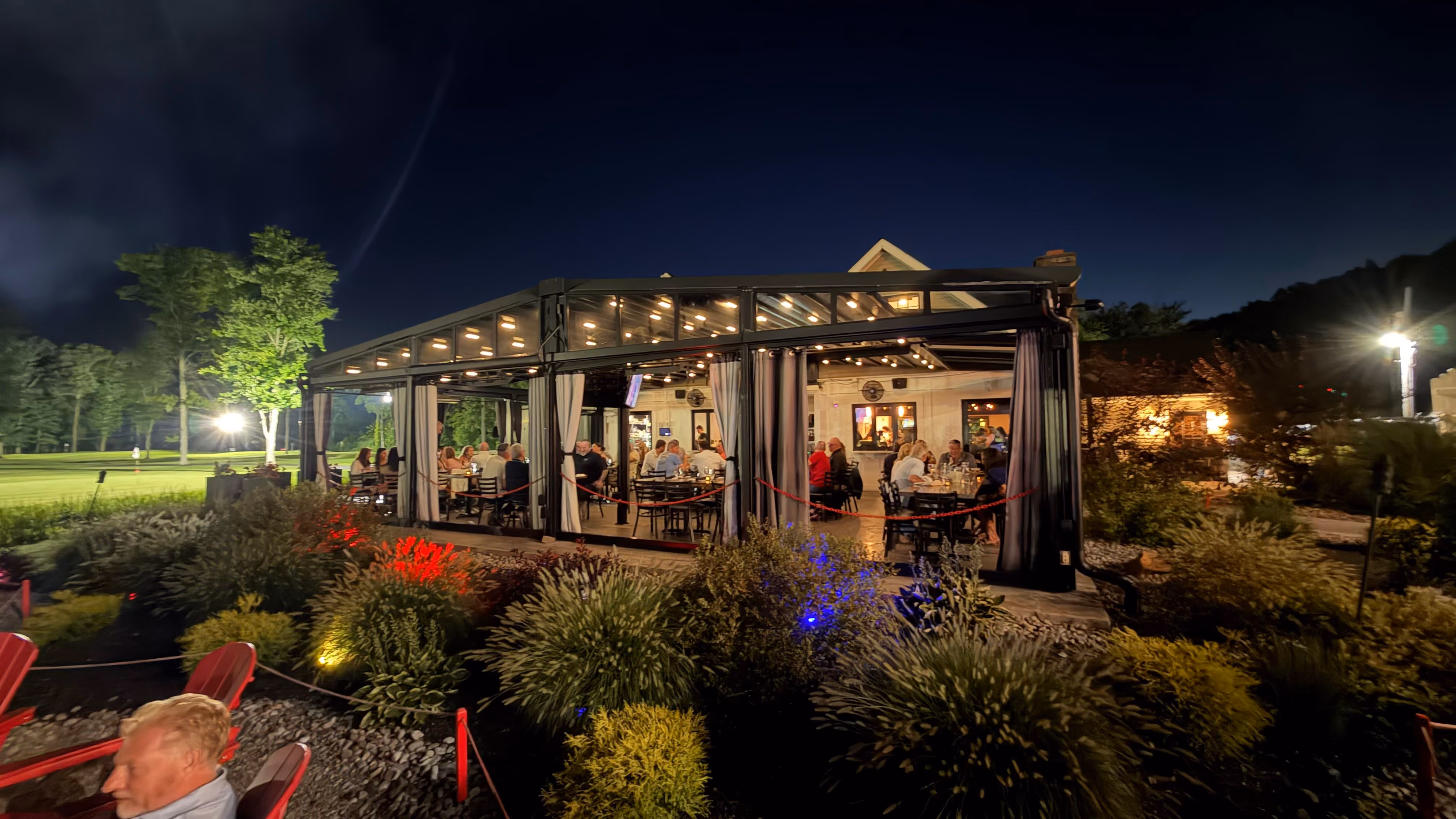 Night scene of people dining inside a glassed patio with garden plants and lawn in the background.