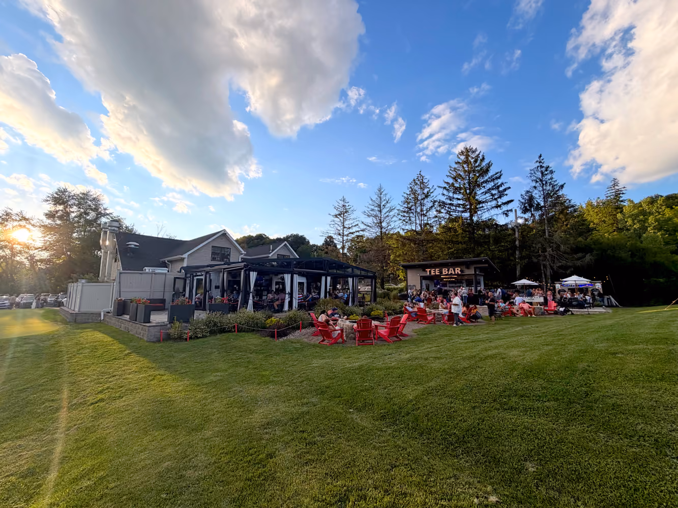 Outdoor bar and lounge area with people sitting in red chairs on a grassy lawn under a partly cloudy sky.