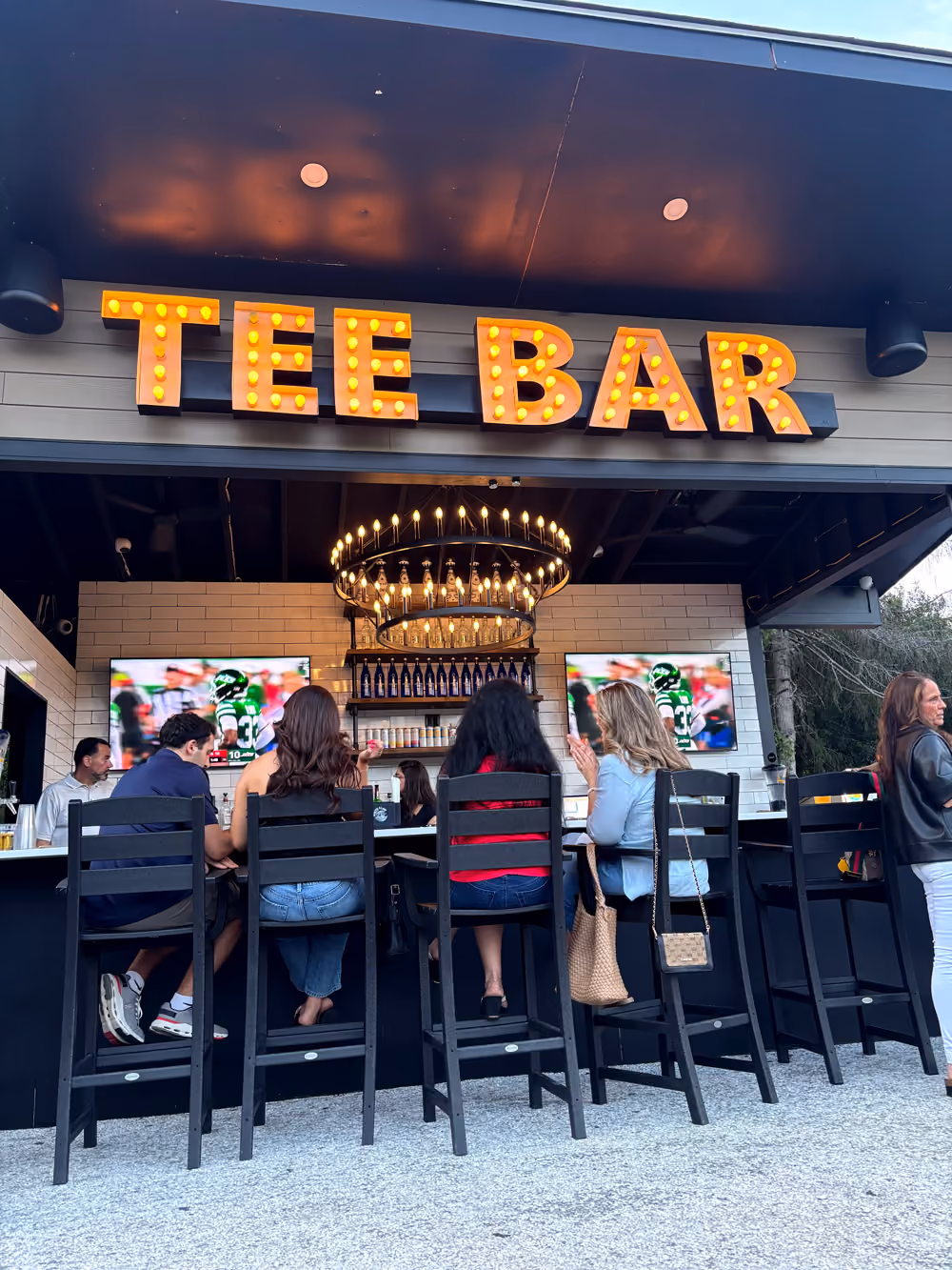 People seated on high chairs at Tee Bar with bright marquee sign and two TV screens showing a football game.