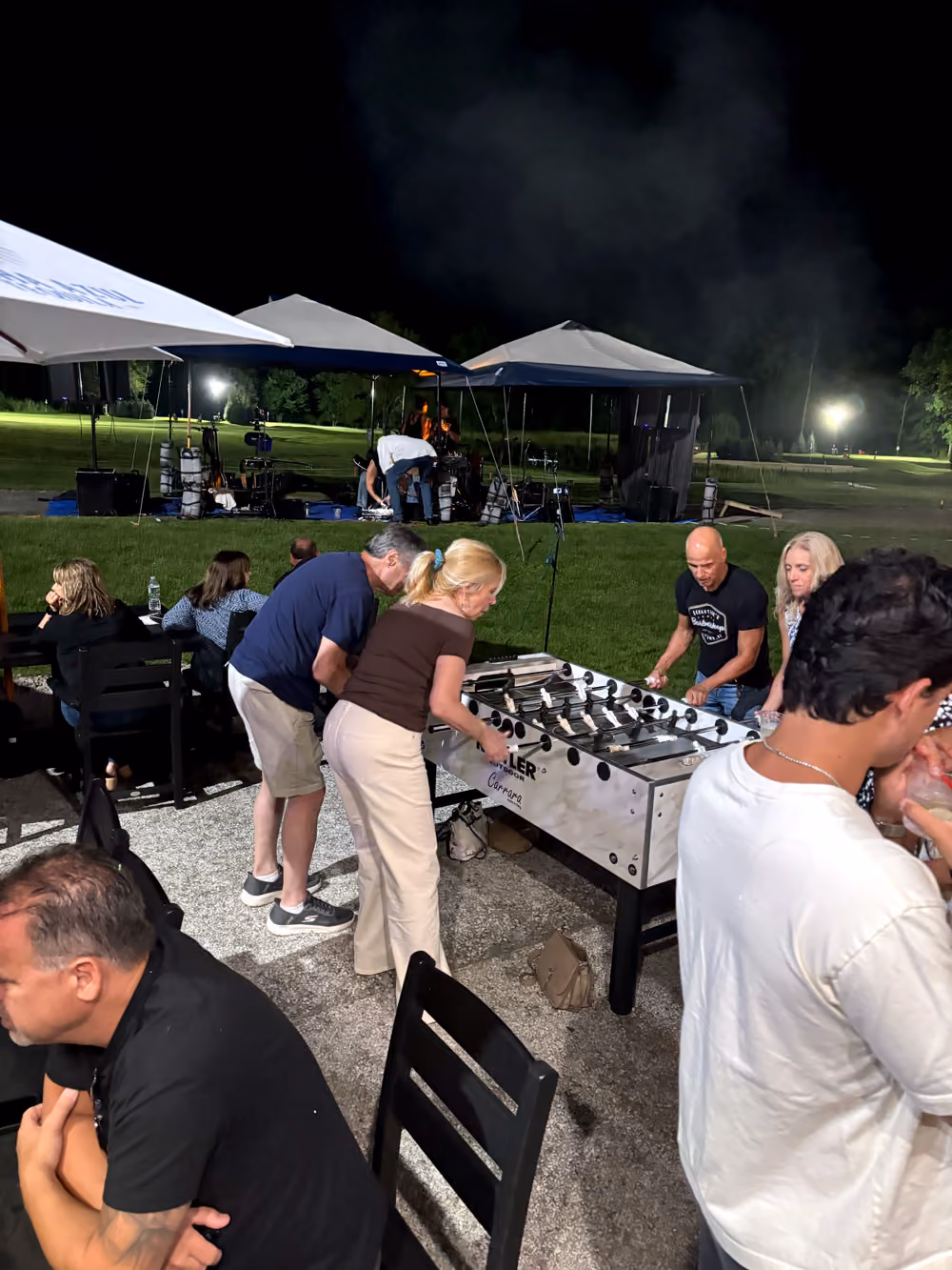 Adults playing foosball outdoors at night with tents and green field in the background.