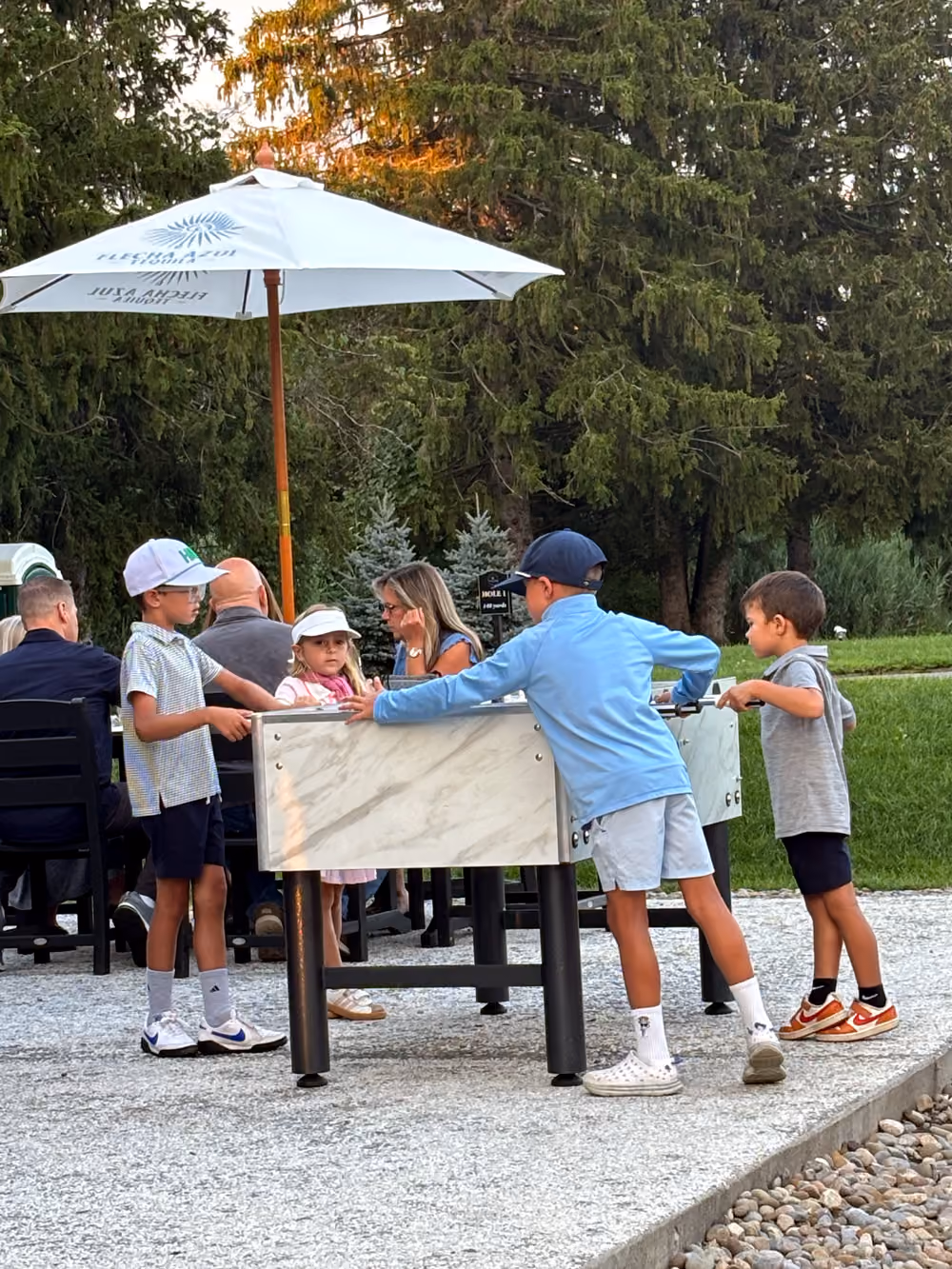 Four children playing foosball outdoors near a white umbrella and adults at a table in a park.
